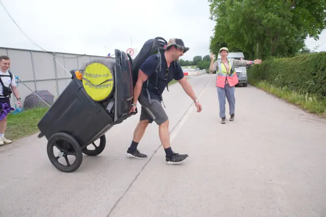 The same man pulls the bin across a tarmac road as he is directed by a steward.