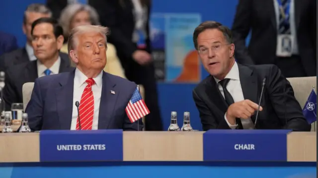 Donald Trump (L) sits down at a wooden circular desk with Mark Rutte (R) sitting to his left. Rutte is leaning towards the US president and appears to be speaking. Diplomats and aides are sitting behind them, including US Secretary of State Marco Rubio