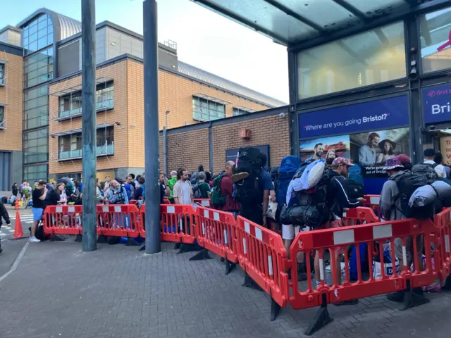 A growing crowd of people carrying large backpacks and luggage queue behind red bollards at Bristol Bus Station.