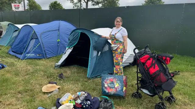 Donna, who wears colourful patchwork dungarees, smiles at the camera in front of her tent. Her belongings are on the ground in front of her and her backpack is in a black pram.