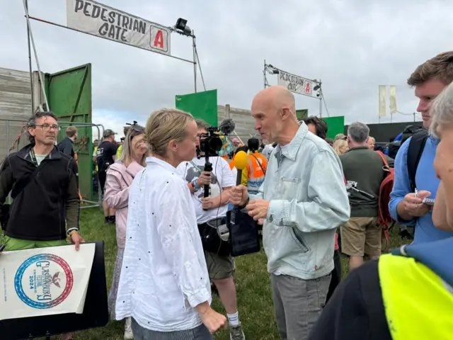 Emily Eavis, who has blonde hair and wears a white shirt, is interviewed by a BBC reporter in front of the main gate. She is surrounded by reporters watching her talk.