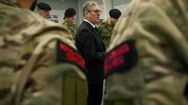 Keir Starmer (C) stands in the centre of a room surrounded by soldiers in green military fatigues talking
