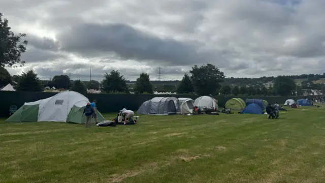 A line of large tents set up in front of a green fence, in a largely-empty field.