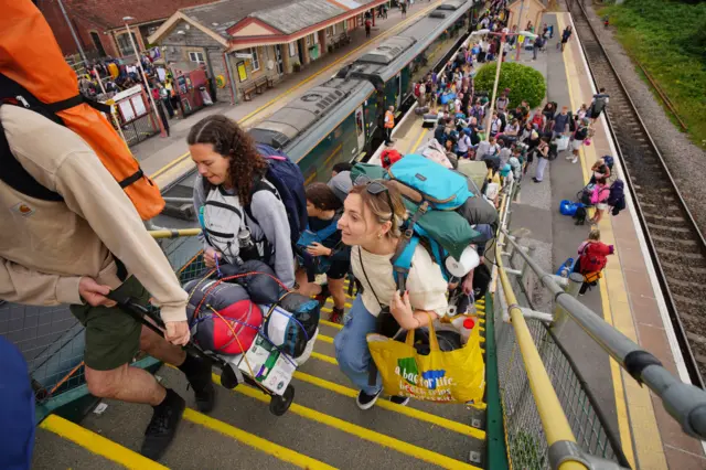 A group of people carrying large backpacks climb the stairs. A train is parked in the station behind them.