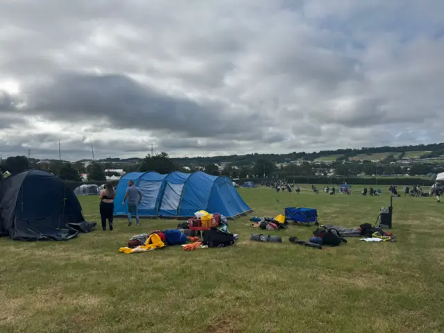 Two large tents are seen the foreground of a camping field, with others visible in the distance.