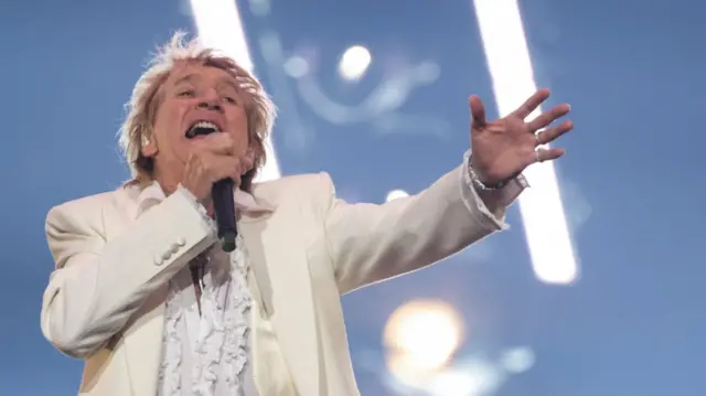 Rod Stewart in white blazer and white ruffled shirt performs on stage, a black mic in his right hand as he extends his left arm in front of him