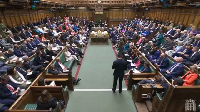Wide shot of House of Commons in session during deputy PMQs, a large group of MPs sitting on the green leather and wood benches, the Speaker visible sitting at the very end of the main central aisle