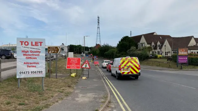 Traffic queuing at contraflow traffic lights on The Strand at Wherstead. A Highways Maintenance van is at the back of the queue in the foreground. The Oyster Reach pub and Premier Inn are on the right of the road. A large To Let sign is on the verge on the left. A brown sign points to Suffolk Leisure Park. A pylon towers on the horizon. The top of the Orwell Bridge is just about visible on the horizon between the pub and the pylon.