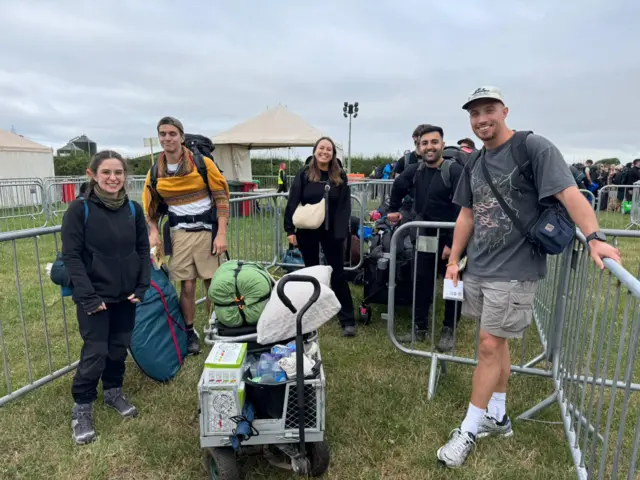 A group of people in outdoors clothing stand between metal railings in a field. They are all smiling at the camera and have large backpacks and trollies full of camping equipment.