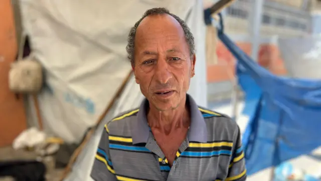 A thin, older man in Gaza looks to the camera. He is wearing a grey collared shirt with yellow, blue and black stripes.