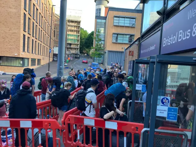 A growing crowd of people carrying large backpacks and luggage queue behind red bollards at Bristol Bus Station.