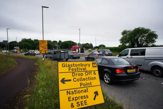 Traffic queueing approaching a roundabout. There is a road sign for Glastonbury Festival in the foreground of the image.