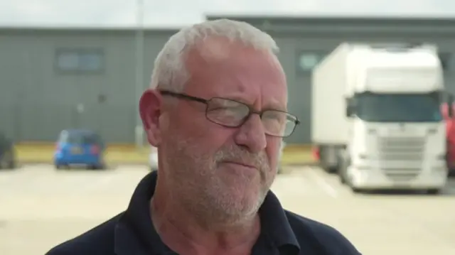 Peter Beard standing in an industrial estate. He is looking away from the camera. He has white hair is wearing glasses as well as a dark polo top.