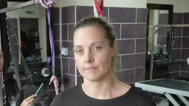 Charlotte McCartney smiles at the camera and has dark hair that has been tied back. She is wearing a black T-shirt. A dog being groomed can be seen behind her.