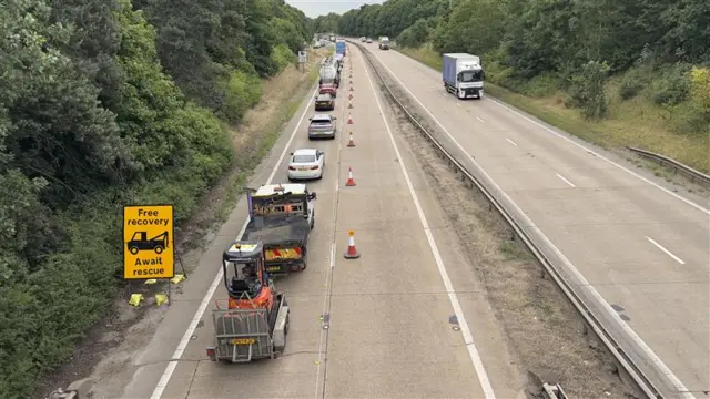 A view of the A14 from over a bridge. The carriageway immediately under the camera has one lane closed off by cones and vehicles are queued up behind one another. The other carriageway is open and a lorry can be seen travelling toward the camera.