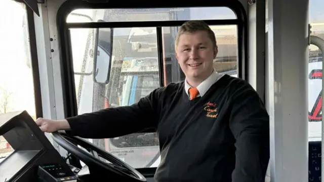 Dan Trent sits in a driver's cab of a coach. He smiles at the camera and has one hand resting on the steering wheel. He has short light hair and wears a jumper with a white shirt and tie underneath.