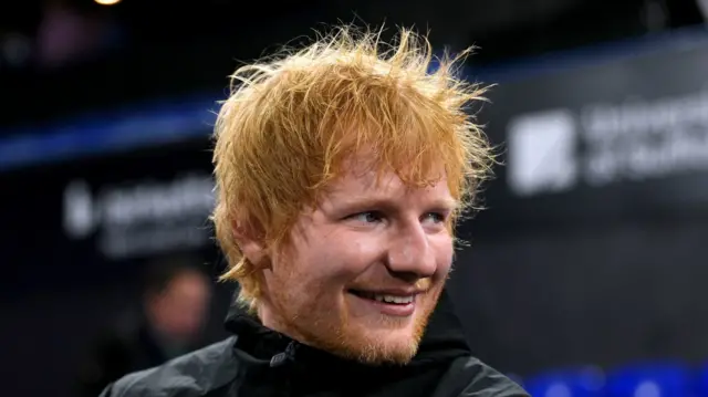 Ed Sheeran is smiling as he looks away from the camera. He is standing in Portman Road stadium. He has shaggy red hair and wears a black waterproof coat.