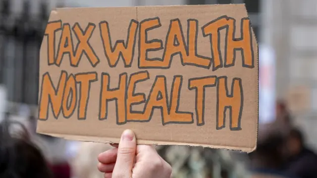 Protest sign reads: "Tax wealth not health"