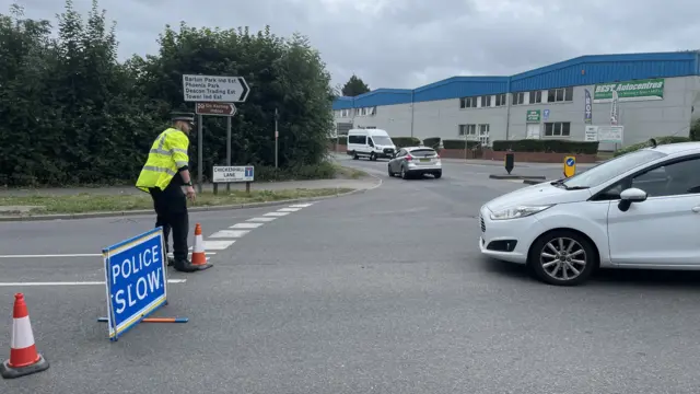 A police officer stands at a "police slow" sign stopping traffic getting near the crash scene