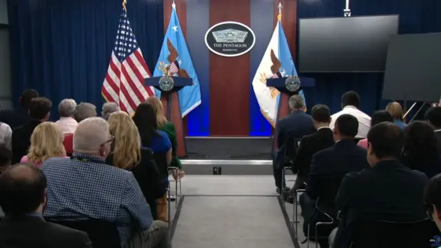 Inside shot of two podiums on stage, DoD flag and US flag in the background, members of the press sitting on metal chairs in the foreground