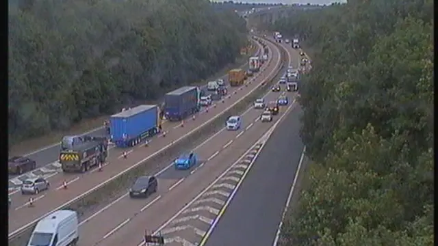 A traffic camera view of the A14 on the approach to Orwell Bridge. One lane is closed on the carriageway further away from the camera and traffic is queuing. The carriageway closest to the camera is open and traffic is moving.