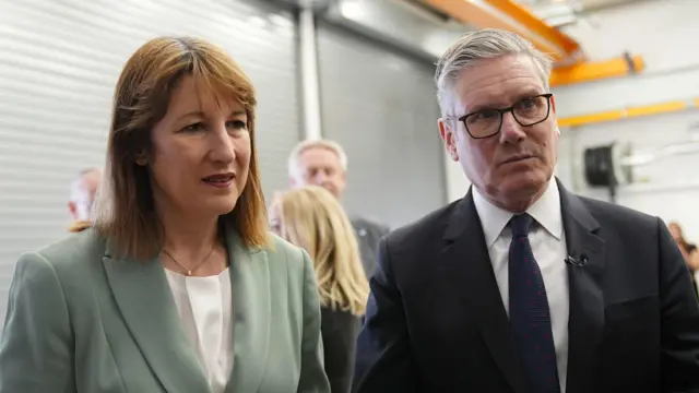 Reeves and Starmer listening to someone in a factory