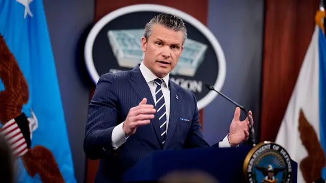 Pete Hegseth in blue suit, white shirt and striped blue tie as he delivers briefing inside Pentagon