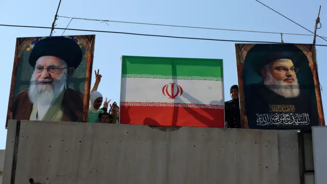 People standing on a rooftop flash the victory sign next to a picture of Iran's Supreme Leader, Ayatollah Ali Khamenei (L), the Iranian flag (C) and a picture of Hezbollah's late leader Hassan Nasrallah.