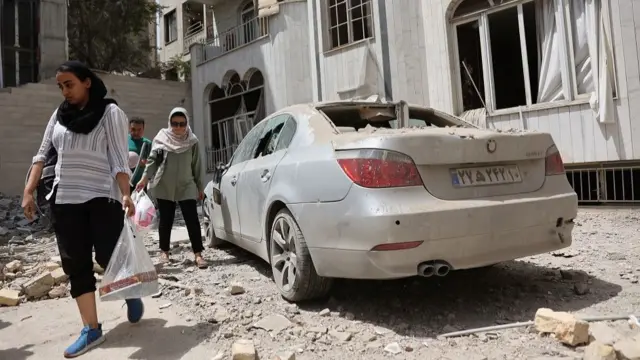 Two women and a man carrying shopping bags walk past a car that is covered in dust and has been damaged in air strikes. There is debris all around them