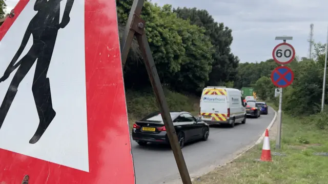 Cars queue to get on the A14. A road sign working of road works is placed next to the road.