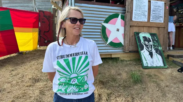Lucinda Tait wears a white and green t-shirt saying Strummerbvlle and looks to the left of the camera, smiling