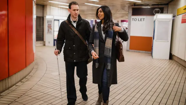 A man with a white stick is guided through a London Underground station