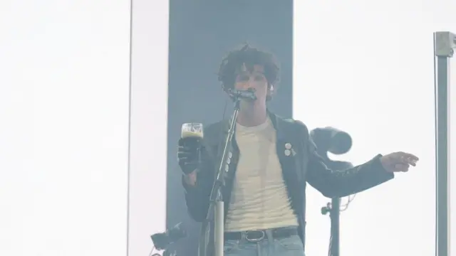 1975'in Matty Healy bir elinde bir bardak bardağı tutup mikrofona şarkı söylüyor