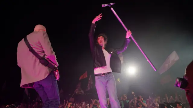 1975 şarkıcısı Matty Healy, kalabalık onun arkasında görülürken mikrofonunu başının üstünde tutuyor