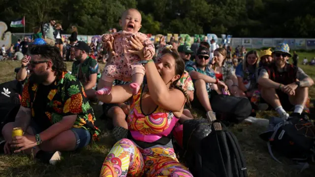 A woman wearing a pink outfit holds up a smiling baby in a small crowd of seated festivalgoers