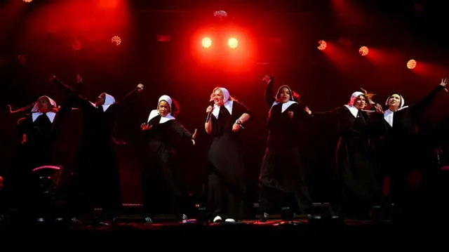 Benlik saygısı siyah bir elbise ve beyaz alışkanlık ile performans gösterir, aynı giymiş yedek dansçılarla dans eder