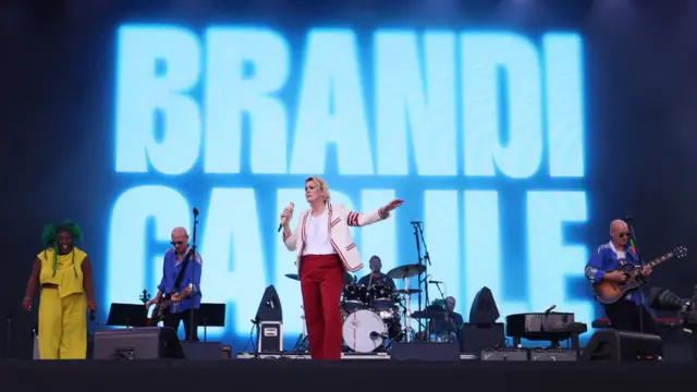 Brandi Carlile performs on the Pyramid Stage with her band. Her name is in big lettering behind her. And she's accompanied by two guitarists, both men wearing blue and sunglasses, and a woman dressed in yellow