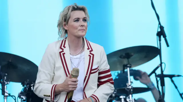 Brandi Carlile wearing a cream jacket with red stripes, and holding a gold microphone as she looks out into the crowd. A drum kit is behind her