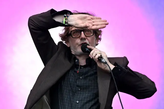 Jarvis Cocker waving his hand on the stage in front of a pink background