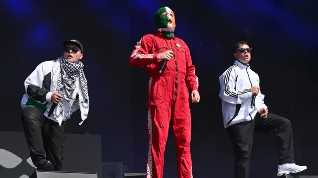 Mo Chara, DJ Provaí and Móglaí Bap of Kneecap. DJ Provai is wearing his tri-colour balaclava and a red boiler suit.