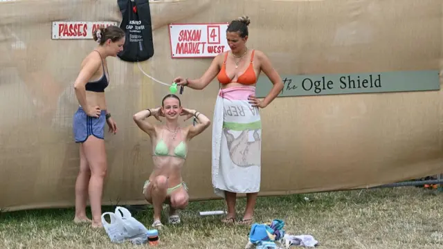 Two women in bikini tops hold up a shower head for another woman, in a green bikini, who is crouching down as she washes