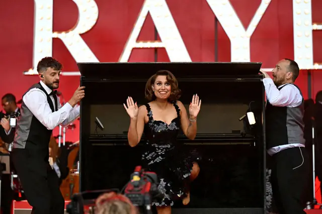 Raye wearing a black sequined dress stood with her hands in the air in front of a piano