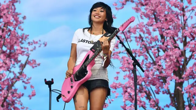 Beabadoobee on stage in front of pink Japanese cherry trees, strumming on a pink electric guitar and wearing a white t-shirt which says 'Glastonbury'