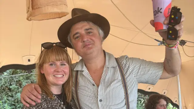 Pete Doherty, wearing a brown hat and striped shirt, holds up a cup as he poses next to BBC reporter Charlotte Gallagher inside a tent, who is smiling