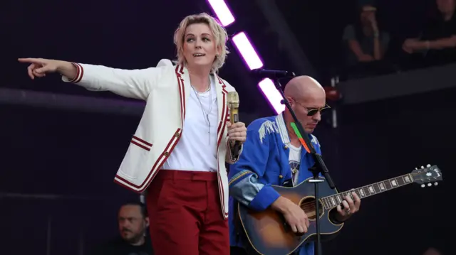Brandi Carlile pointing to the right of the stage, off camera. She's wearing red trousers and a cream jacket and is standing next to an acoustic guitarist wearing a blue jacket