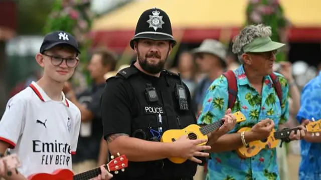 A police officer joins a boy and a man in a group ukulele performance near to the Circus Field