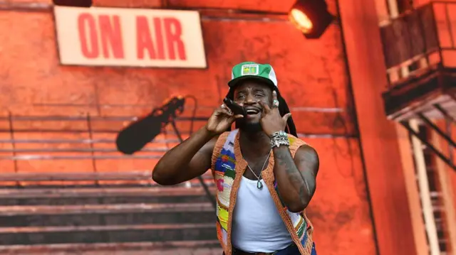 wearing a green and white cap, and a multicoloured knitted waistcoat on top of a white vest. he is wearing navy jeans. he is holding the mic to his mouth with his right hand and is gesturing to his face with his left hand. The backdrop is an orange and looks to be an industrial type building. the words on aii and a mic are also on it