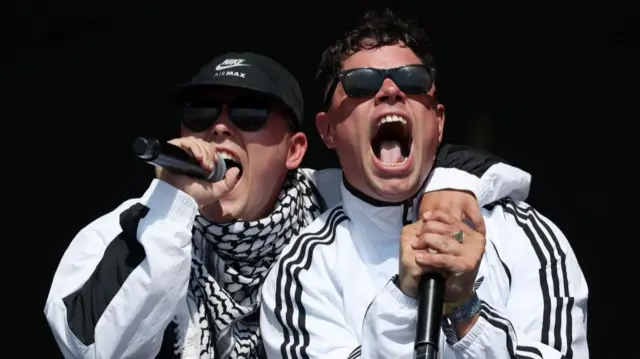 Kneecap's Mo Chara (L) and Moglai Bap (R), dressed in black and white perform animatedly into microphones