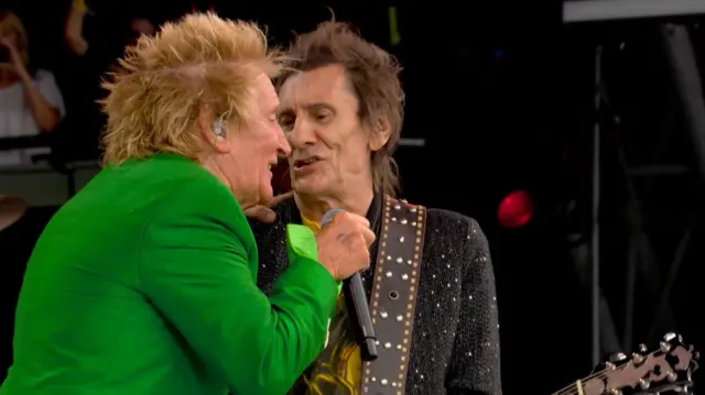 Rod Stewart, wearing a bright green suit, and Ronnie Wood sing into the same microphone