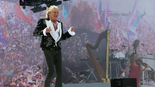 Rod Stewart, wearing a black jacket with gold details and stripy pants, dances on stage